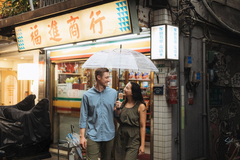Couple sharing umbrella in rain outside Asian shop with Chinese characters on sign.