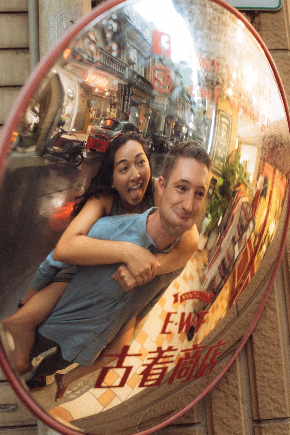 Couple smiling and posing together reflected in a convex street mirror in an Asian city alleyway.