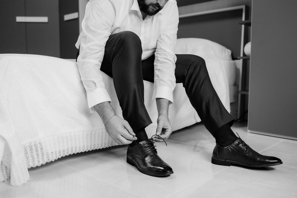 Lo sposo si allaccia le scarpe seduto sul letto, in bianco e nero.