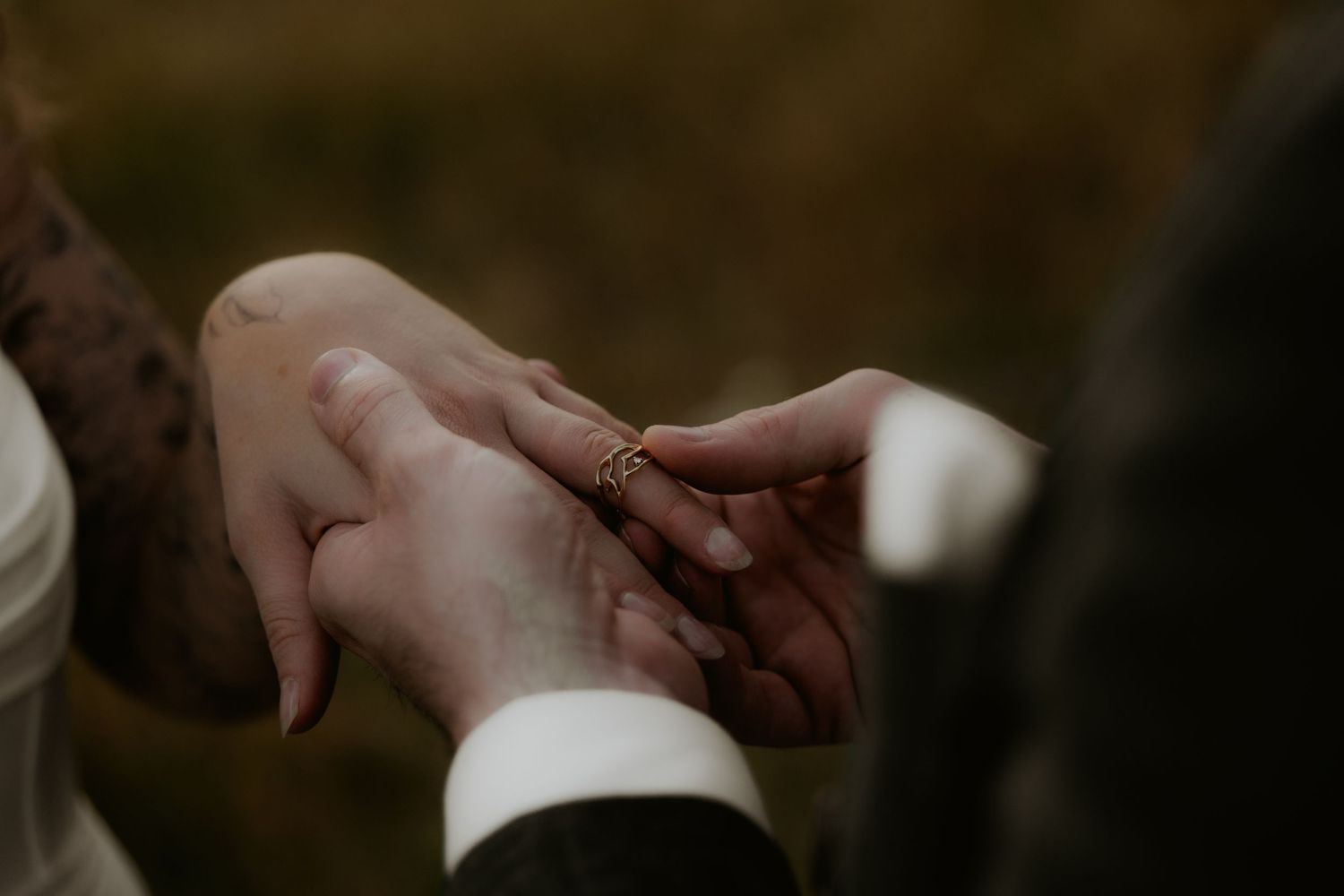 A groom places a gold wedding ring on the bride's finger during an outdoor wedding ceremony.