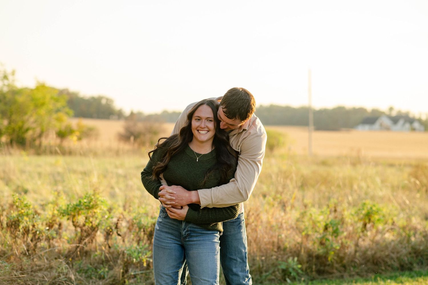 Man hugs laughing woman from behind during romantic golden hour engagement session in autumn field.