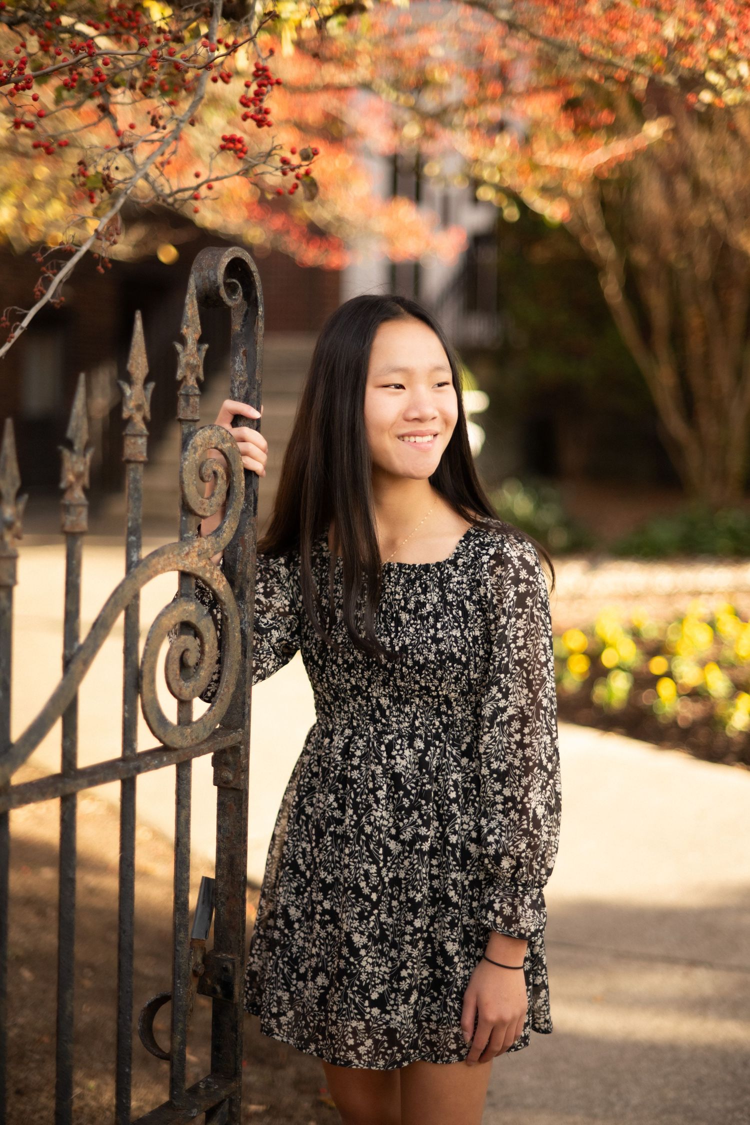 Young Asian woman in floral dress poses by ornate iron gate with autumn foliage in background.