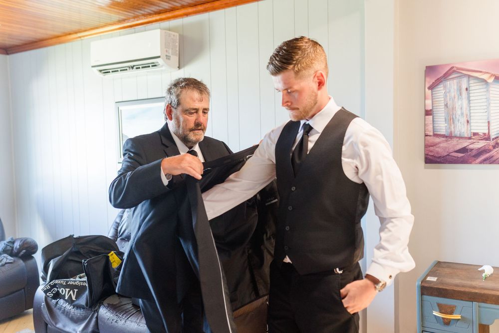 Two men in formal suits prepare for a wedding, with one helping the other put on a suit jacket indoors.