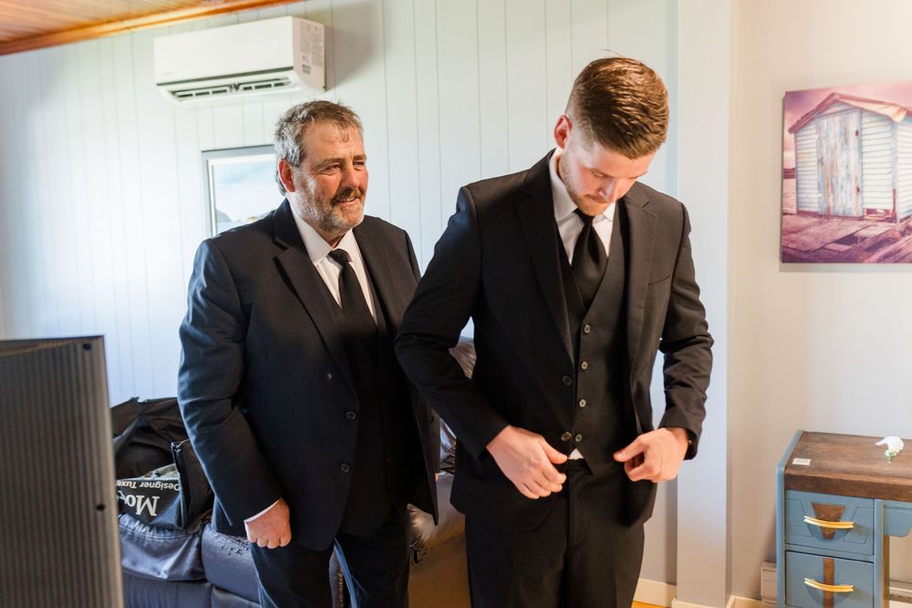 Two men in black suits adjusting attire indoors, preparing for a formal wedding ceremony.