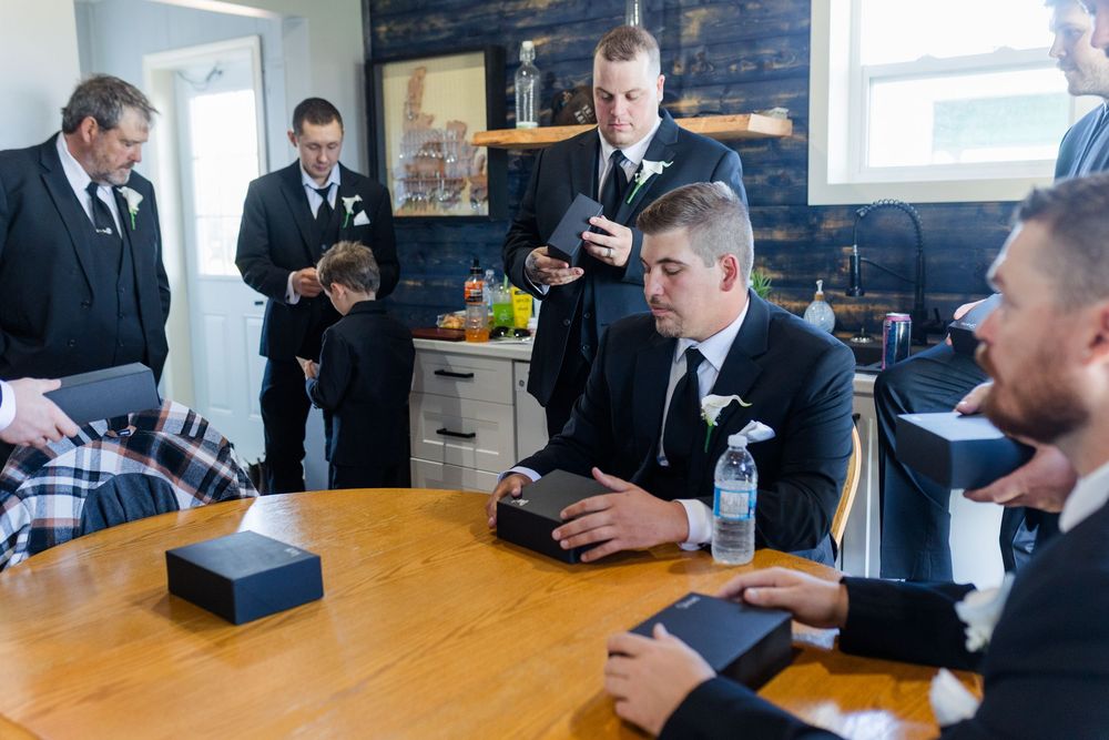 Groomsmen in black suits gather around a wooden table, opening gift boxes before a wedding ceremony.