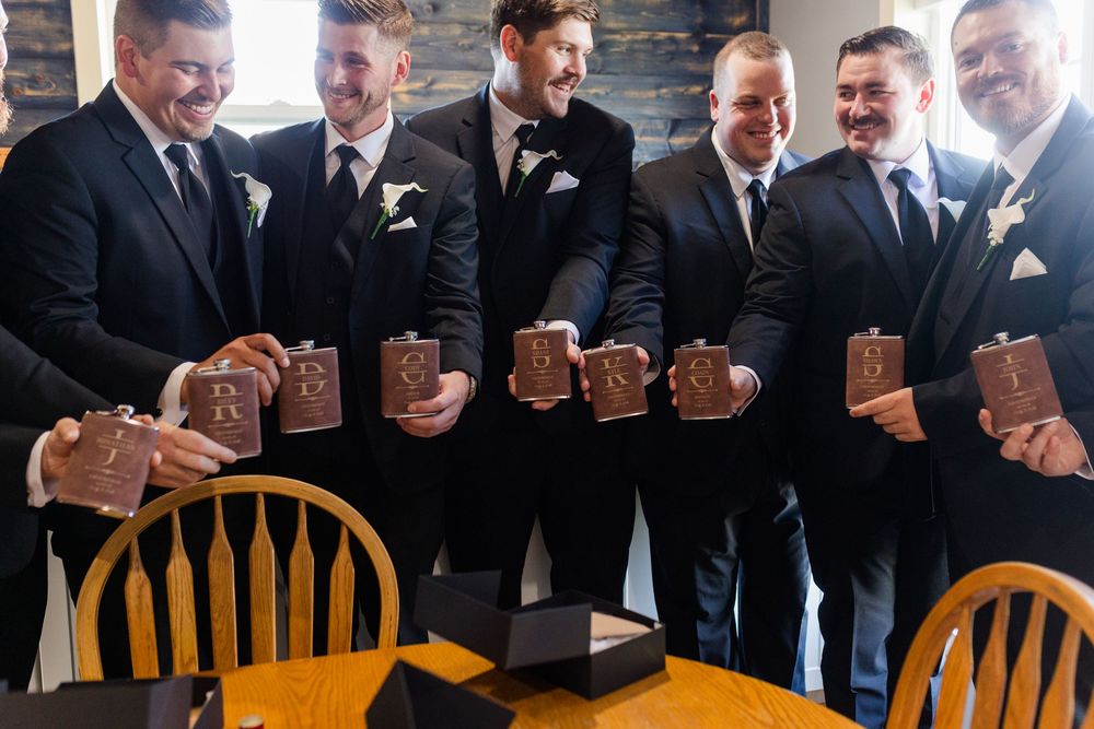 Groomsmen in black suits smiling and holding engraved wooden flasks at a wedding celebration.