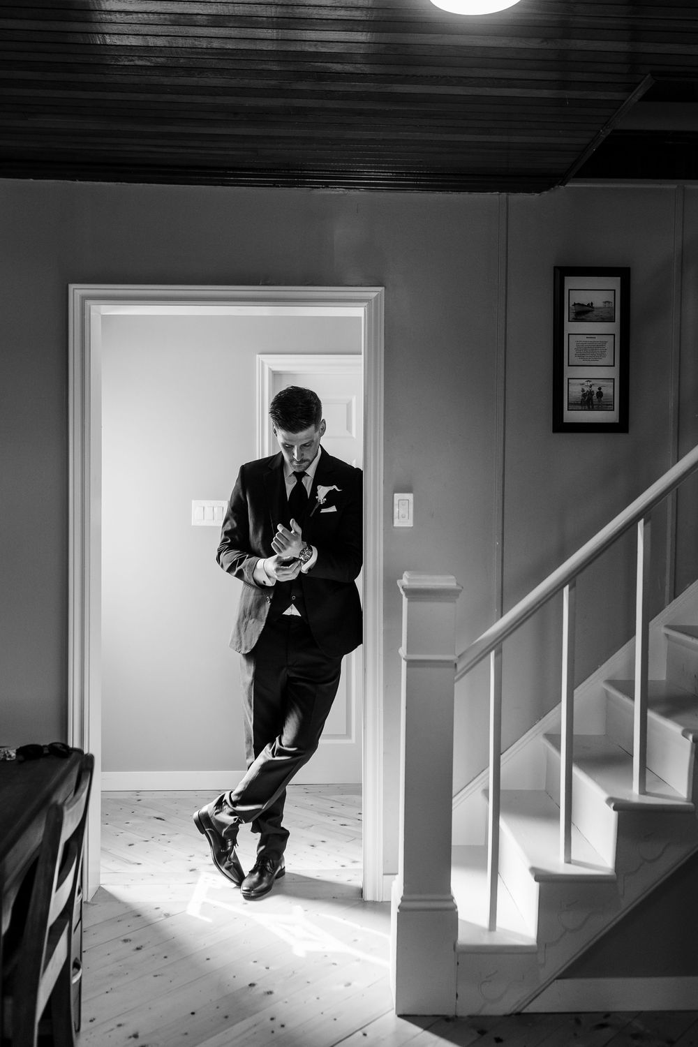 Groom in black suit adjusting cufflinks in doorway, black and white wedding photo with staircase.