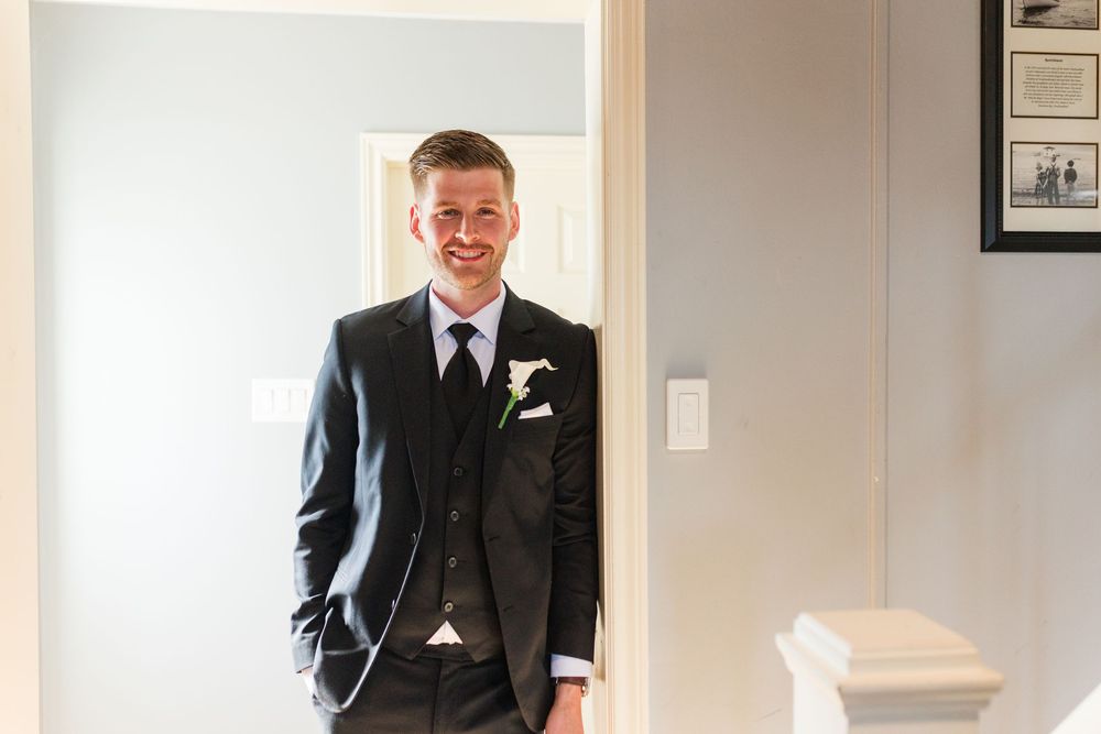 Smiling groom in dark suit with boutonniere stands in doorway on wedding day.