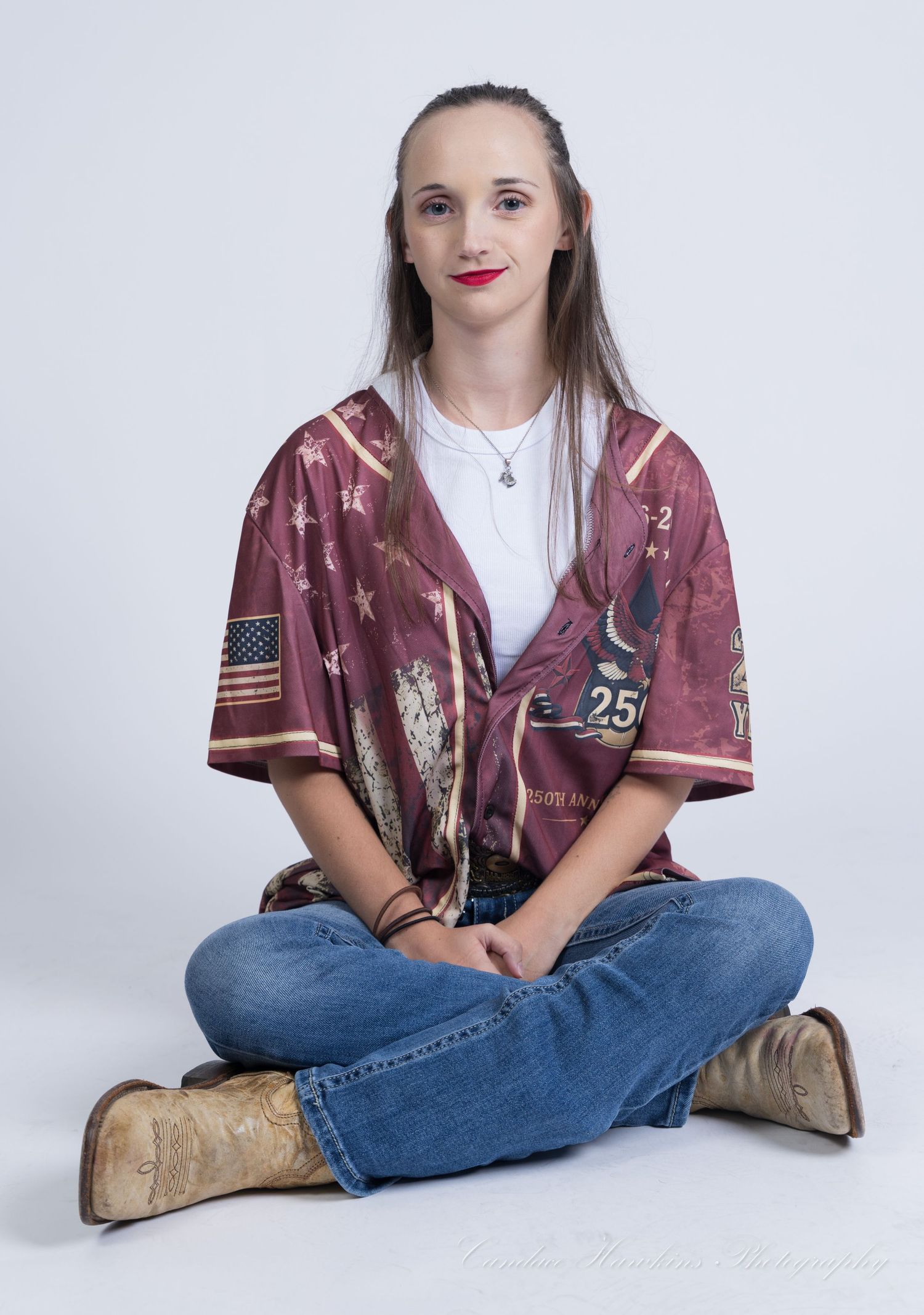 Young woman with long brown hair and red lipstick sits cross-legged in a maroon baseball jersey, jeans and tan cowboy boots.