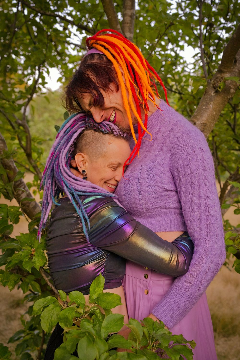 Two women with colorful dreadlocks embrace and laugh joyfully outdoors among green trees.