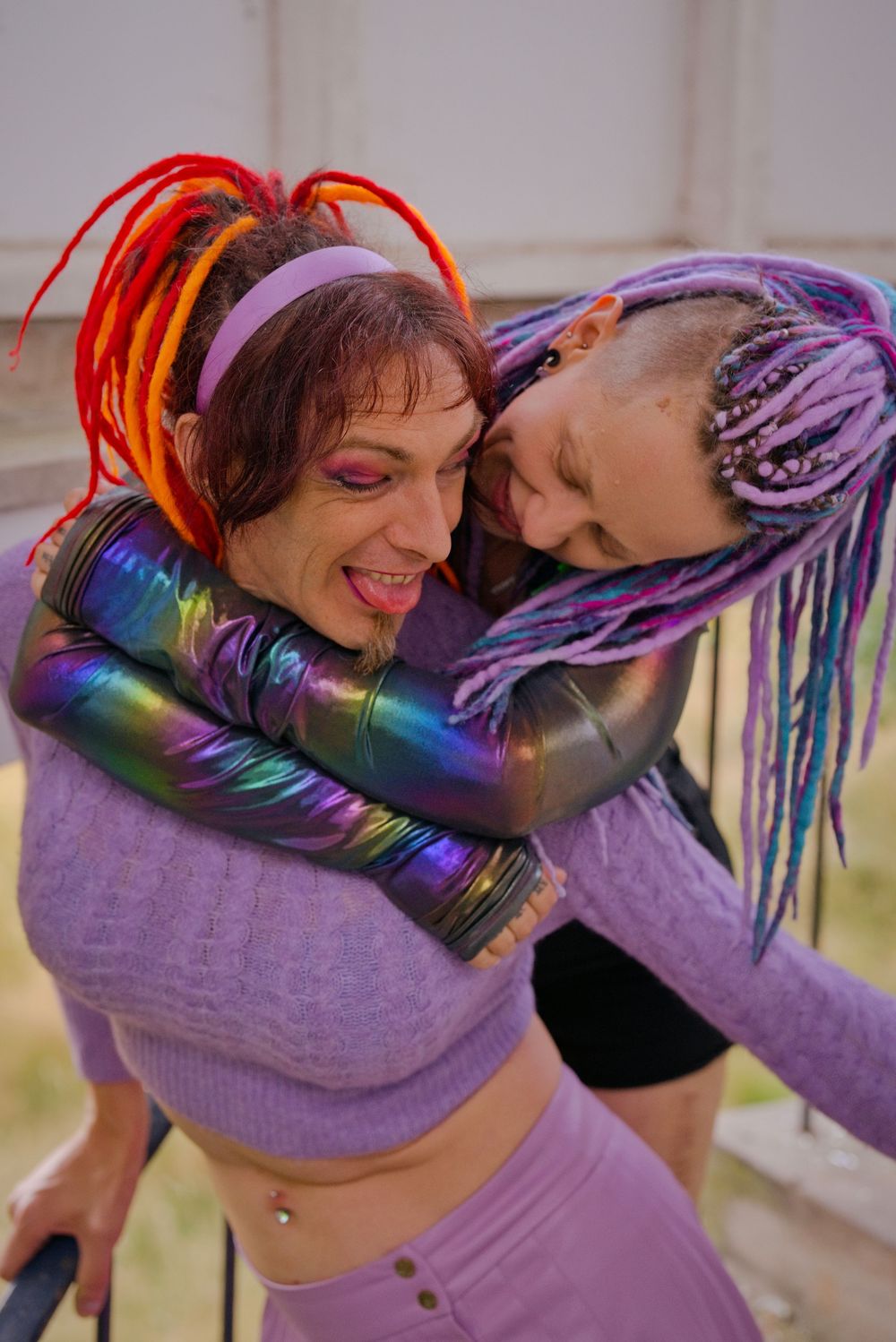 Two people with colorful dreadlocks in purple and rainbow hues embrace playfully outdoors wearing vibrant festival outfits.