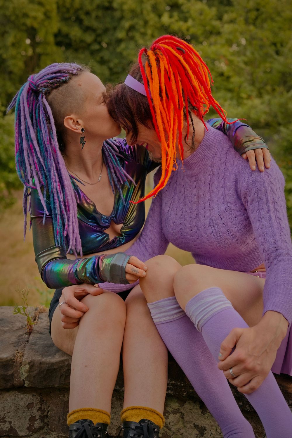 Two women with colorful dreadlocks sharing a tender moment outdoors, wearing vibrant purple and holographic outfits.