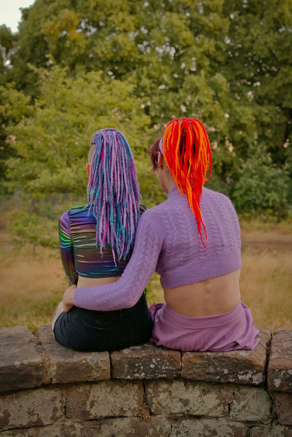 Two women with colorful dreadlocks sit together on a stone wall, embracing outdoors.