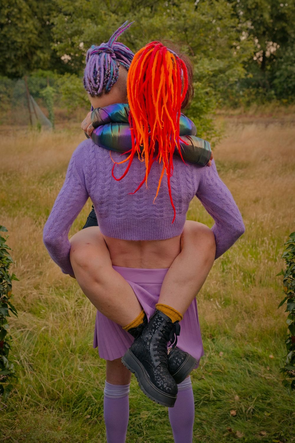 Person with orange dreadlocks carried piggyback by someone in purple sweater and skirt outdoors.