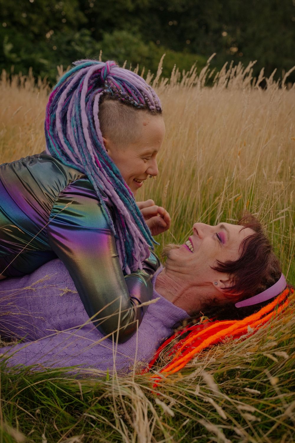 Two people in colorful outfits laugh together while lying in a golden grass field outdoors.