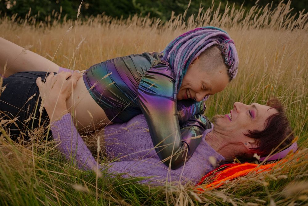 Two people sharing a tender moment lying in a golden grass field, one wearing a colorful hijab and iridescent top.