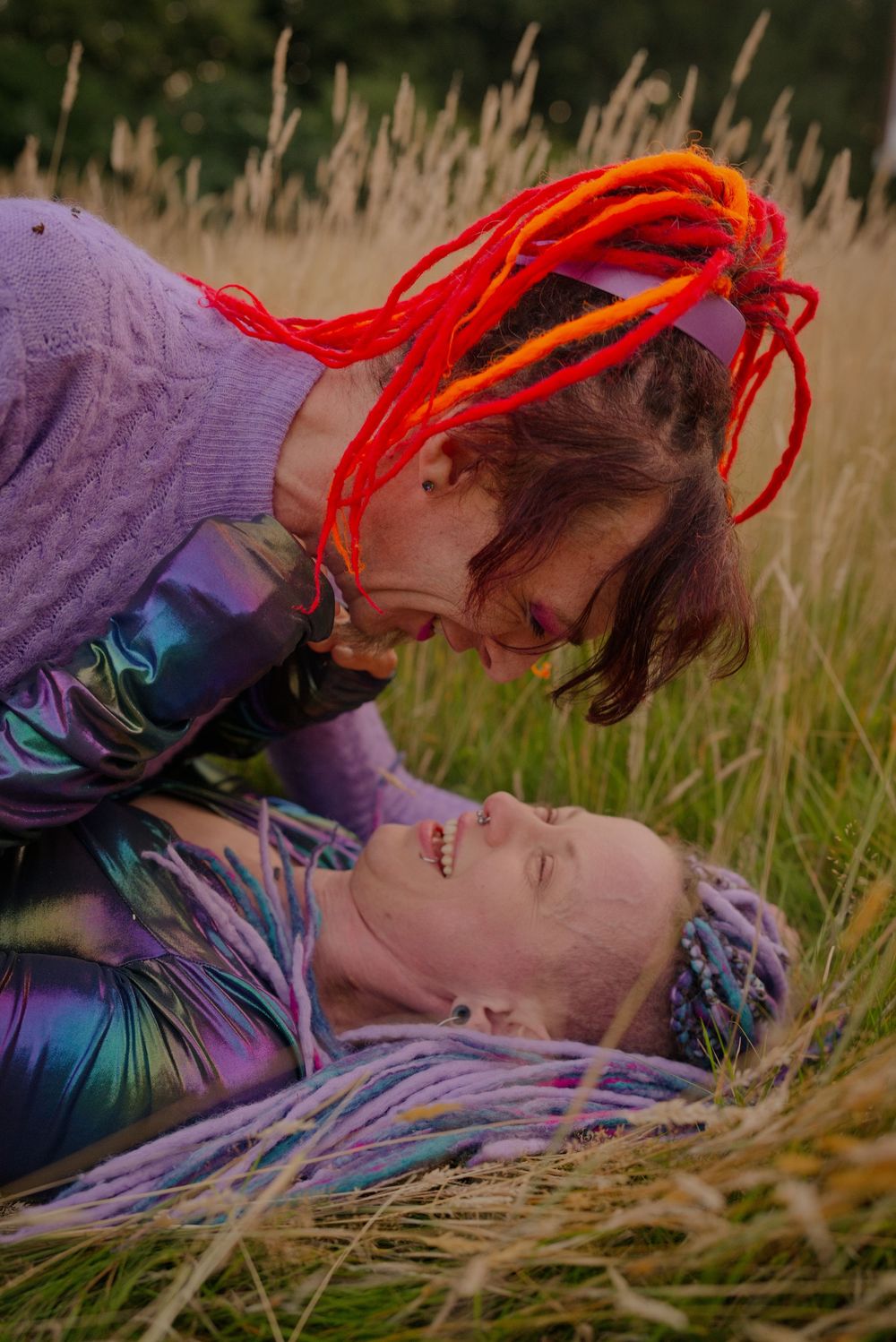 Two people with colorful dreadlocks share an intimate moment lying in a golden grass field outdoors.