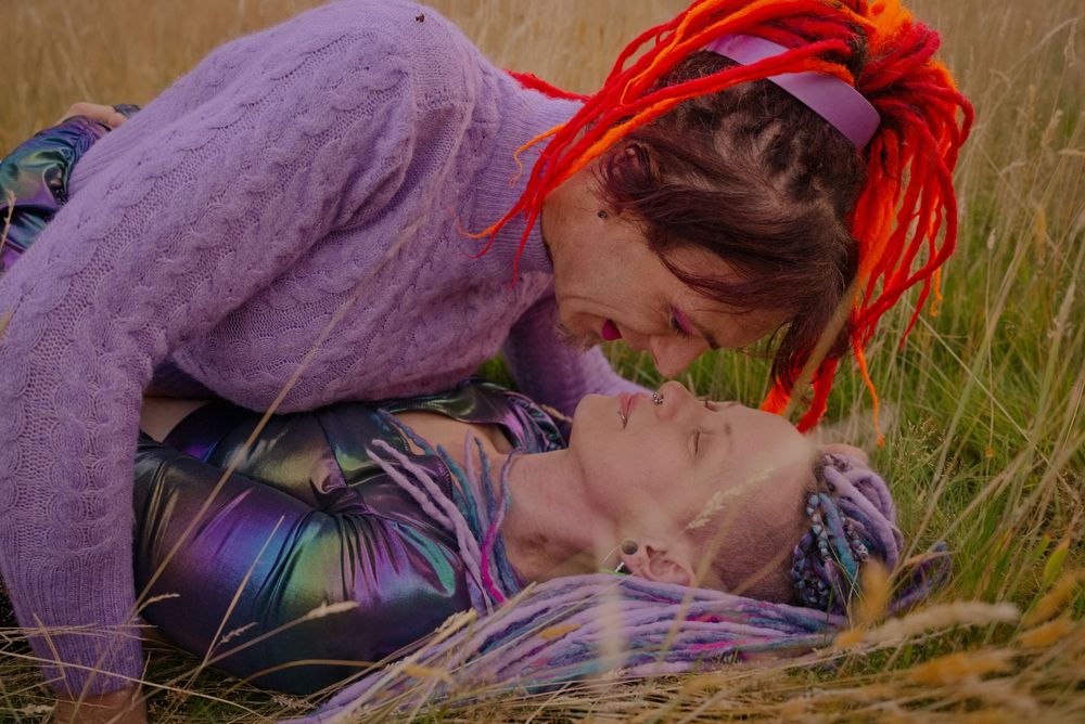 Two people sharing a romantic moment lying in a golden grass field, one with vibrant red dreadlocks.