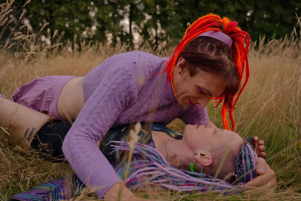 Two people with colorful hair share a romantic moment lying in a golden grass field outdoors.