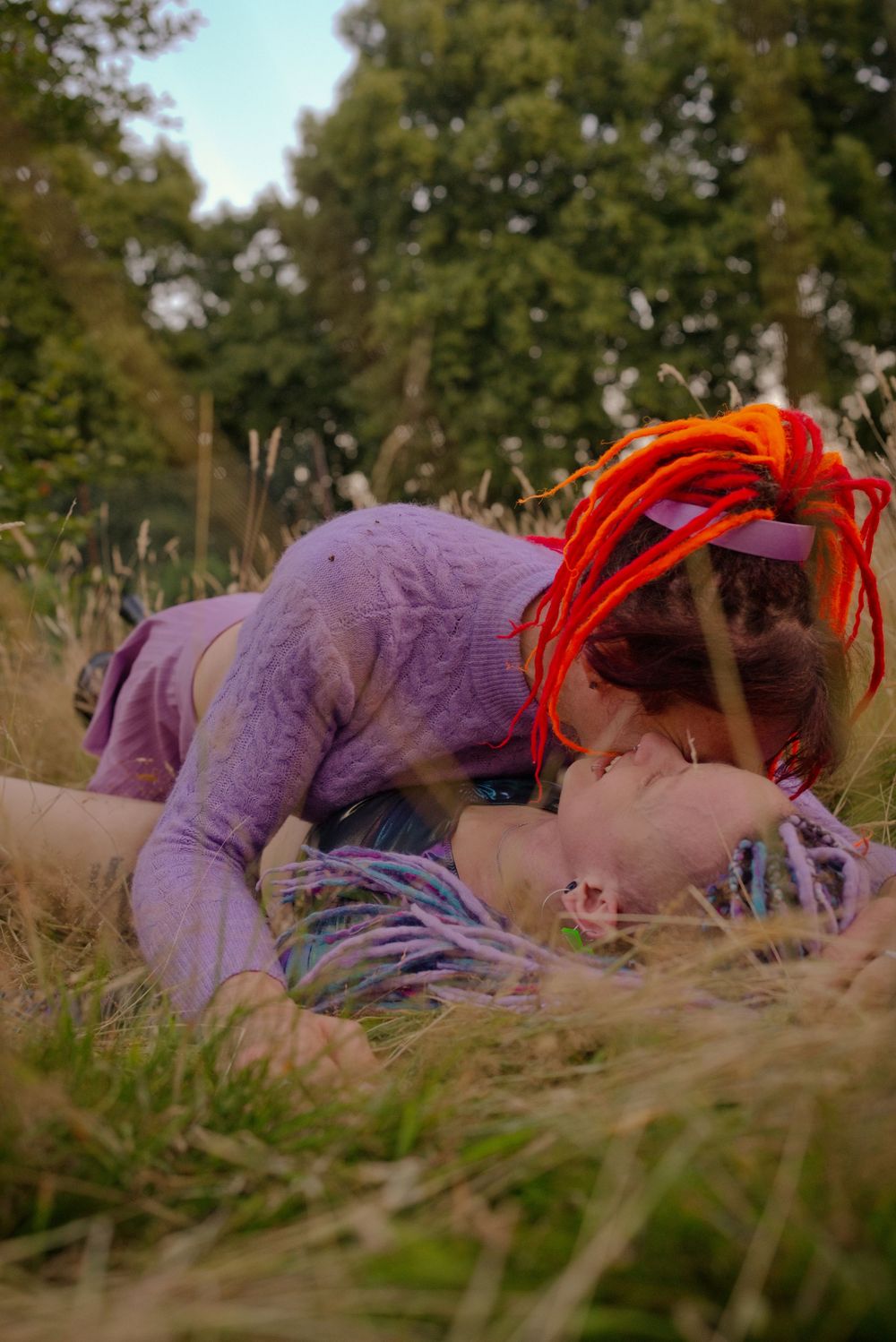 Two people with colorful dreadlocks lying in tall grass outdoors near trees.