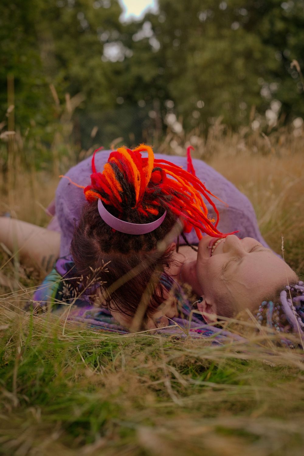 Woman with vibrant red and orange dreadlocks lying in tall grass wearing a purple headband and floral dress.