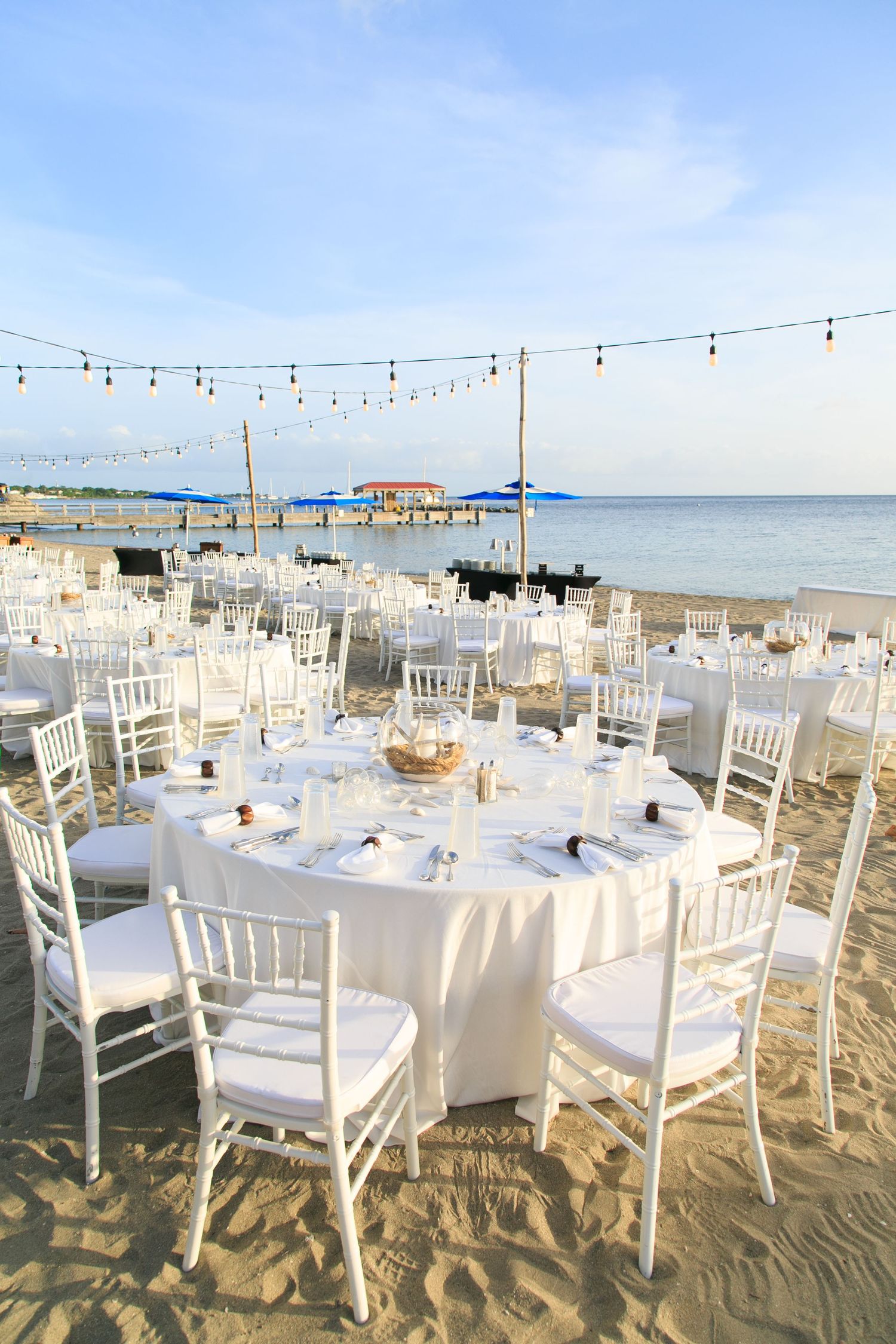 Elegant beach wedding reception setup with white round tables, chiavari chairs, string lights, and ocean views at sunset.