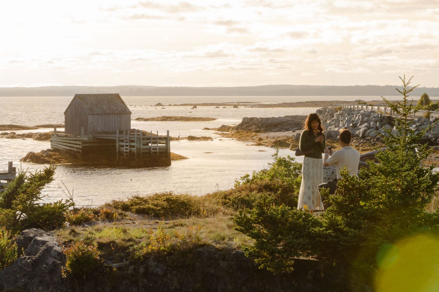 Man and woman stand on rocky coastal cliffs at golden hour, with a rustic fishing shack and calm ocean waters in the background proposing.