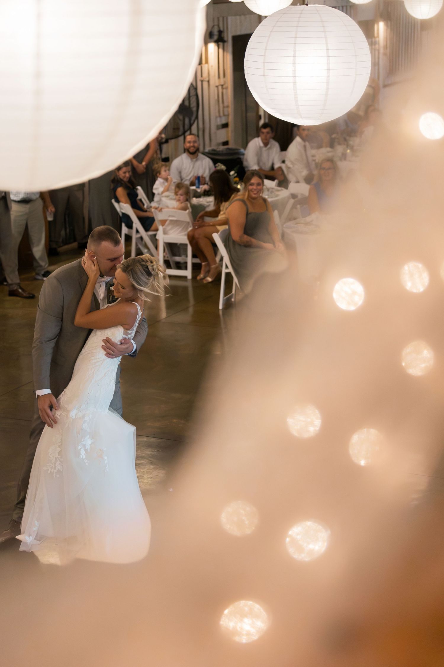 Rustic barn wedding reception with white round paper lanterns hanging from wooden ceiling above elegantly set round tables.