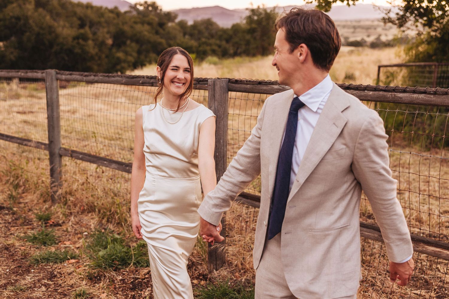 Couple holding hands walking by a rustic fence in a golden field, woman in satin dress, man in beige suit.