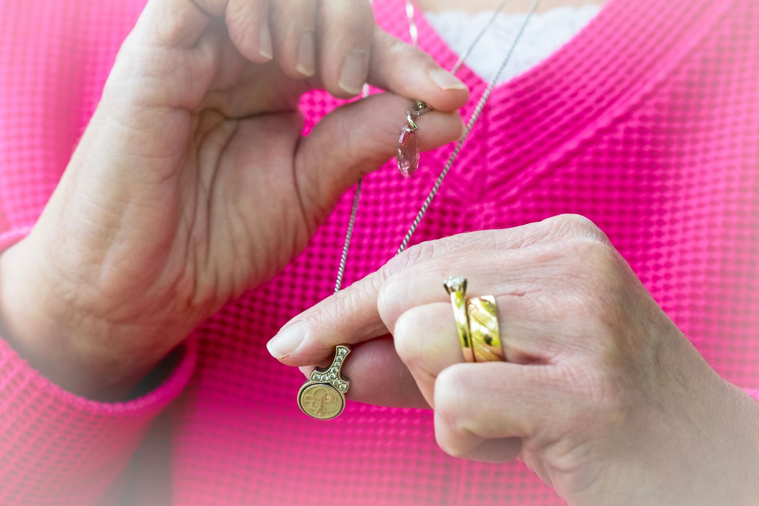 Woman in pink top fastening a delicate gold chain necklace with pendant charms, wearing a gold ring.