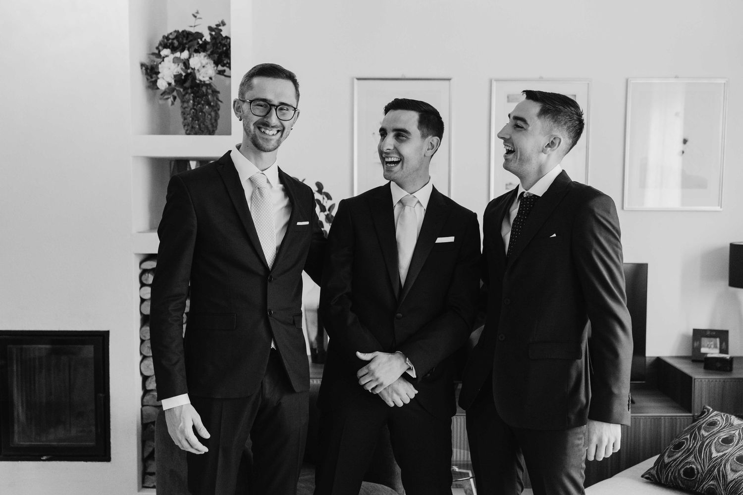 Three men in suits laughing together at a wedding celebration in a bright indoor setting.