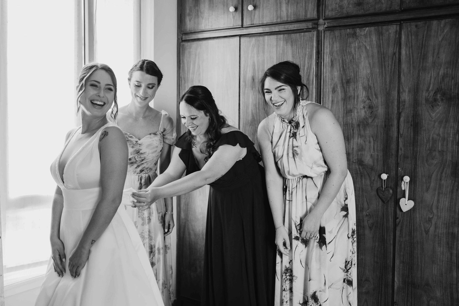 Black and white photo of a bride laughing with three bridesmaids while getting ready near wooden doors.