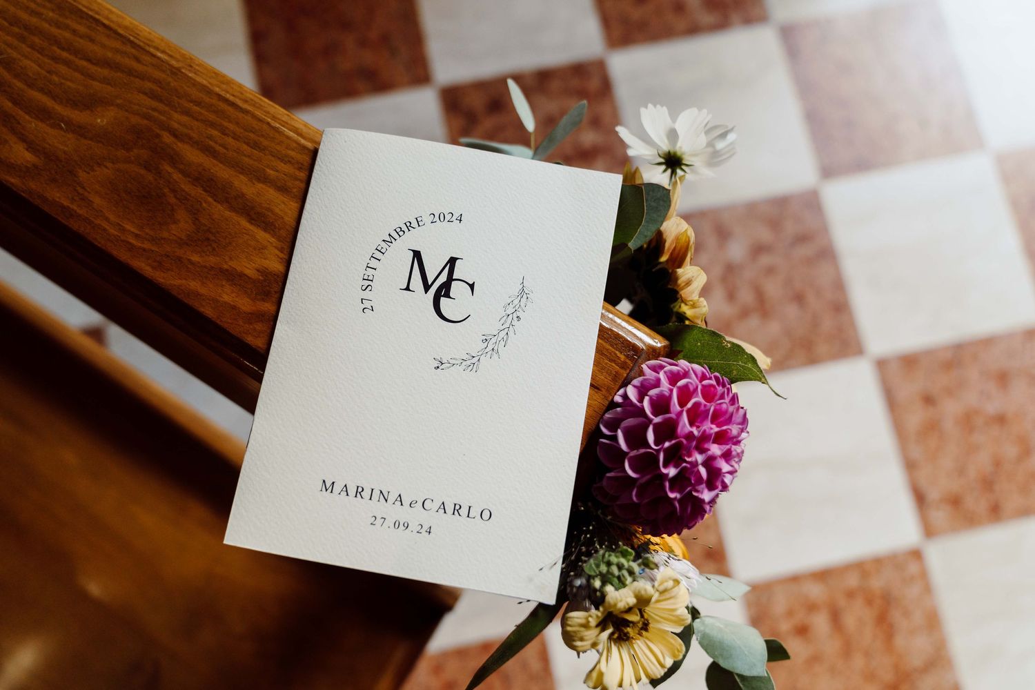 Elegant wedding program for Marina and Carlo dated 27 September 2024, displayed with colorful flowers on a checkered floor.