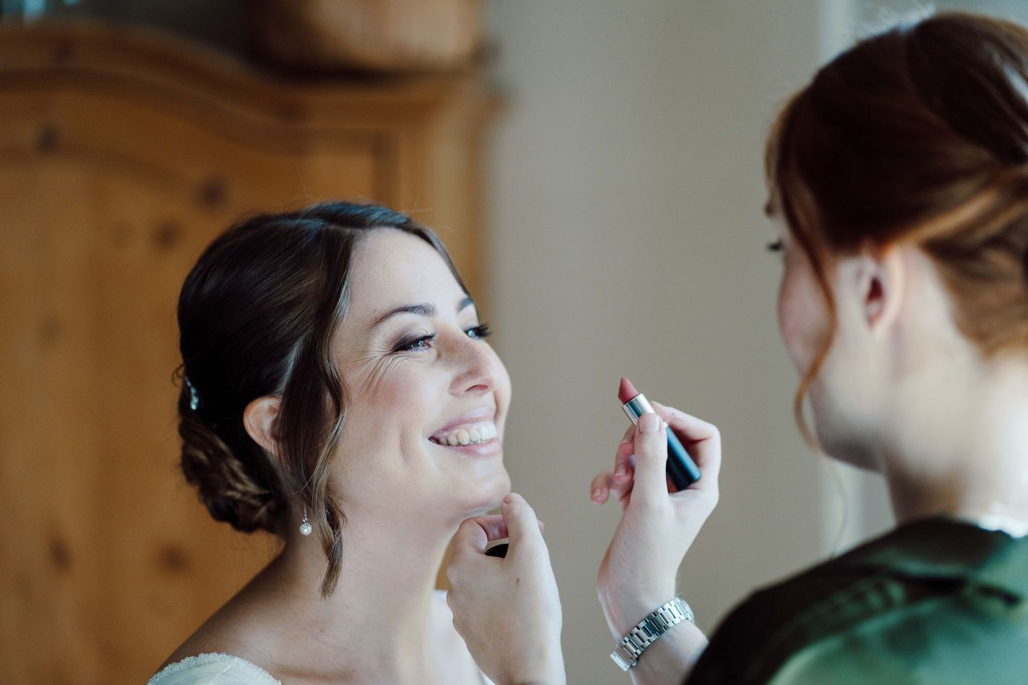 Makeup artist applying lipstick to a smiling bride during wedding preparations.