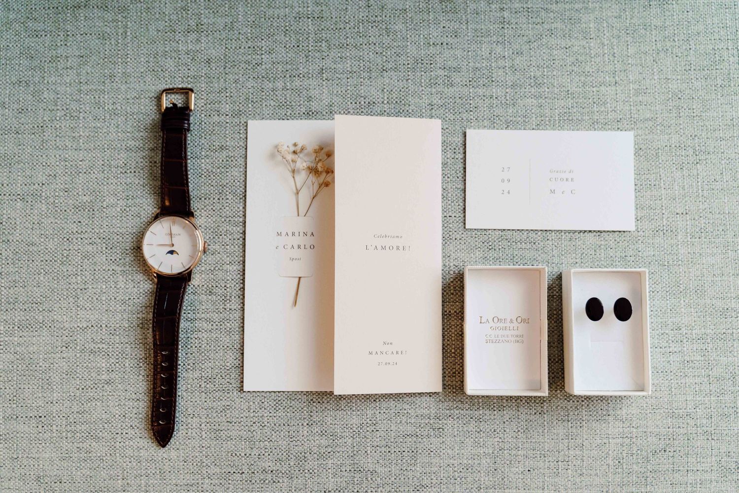 Flat lay of wedding stationery, a classic watch, dried flowers, and elegant stud earrings on a neutral linen background.