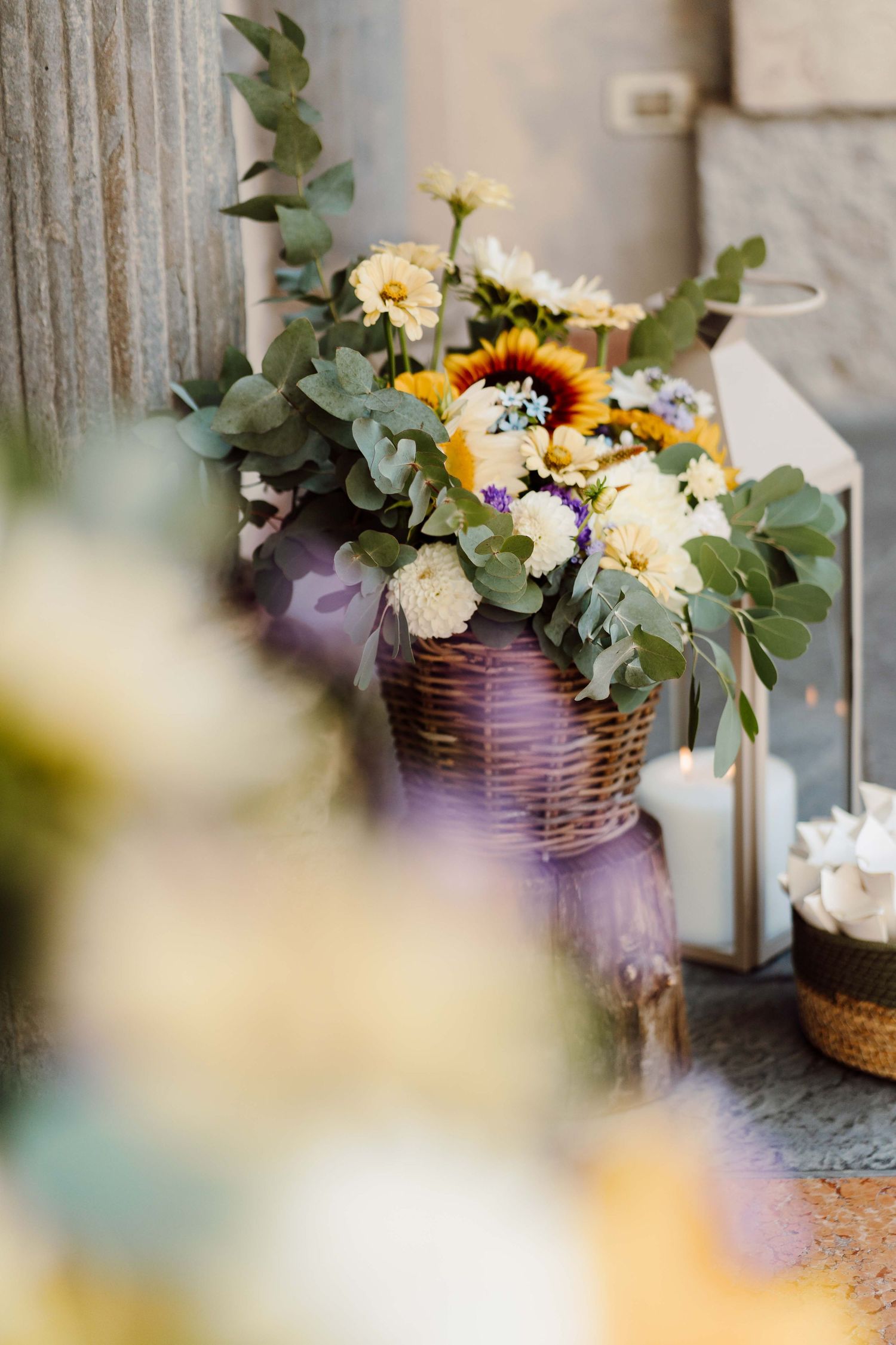 Rustic wicker basket with yellow, white, and red flowers and eucalyptus beside a candle lantern at a wedding.