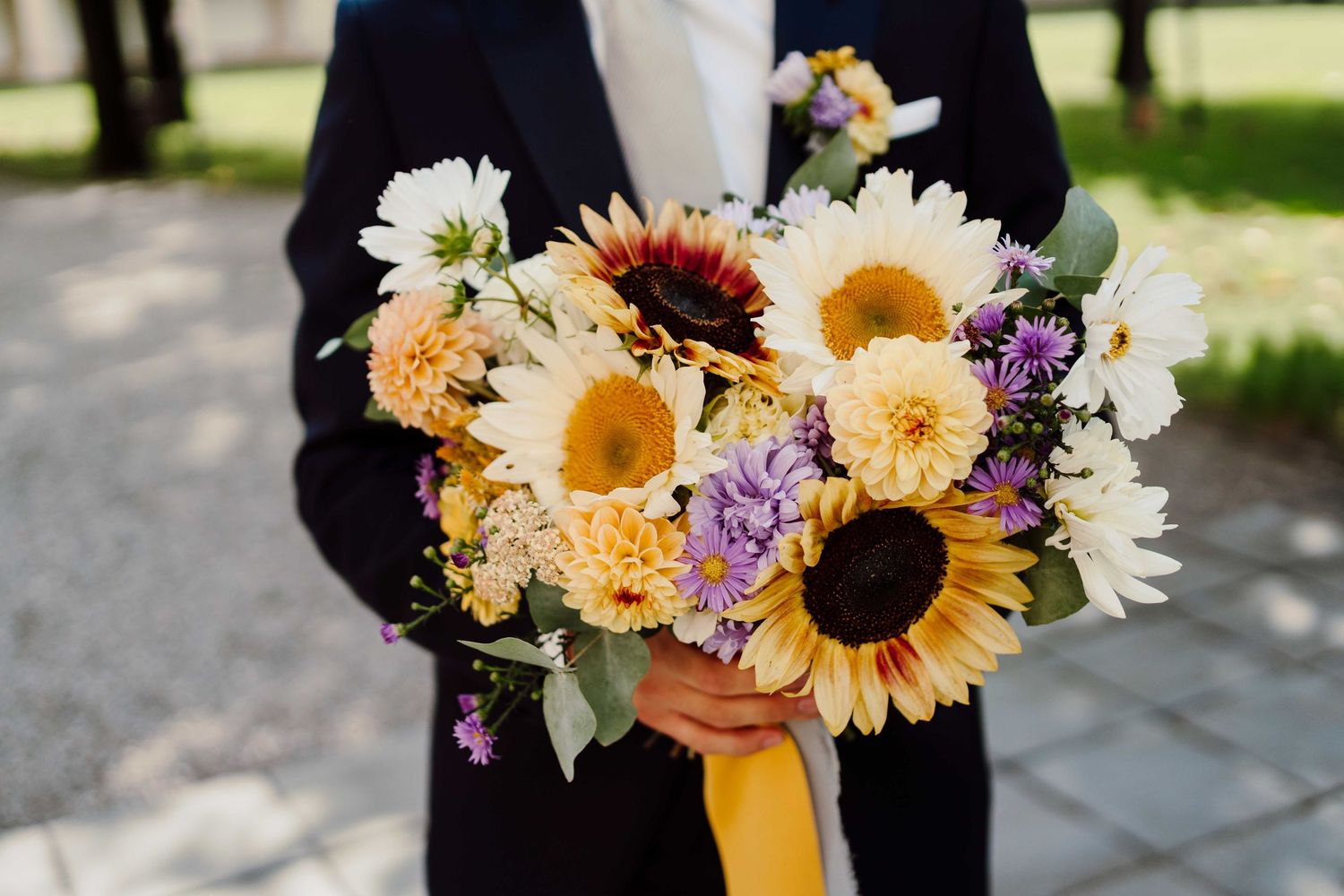 Person in navy suit holding a vibrant wedding bouquet with sunflowers, white daisies, and purple wildflowers tied with yellow ribbon.