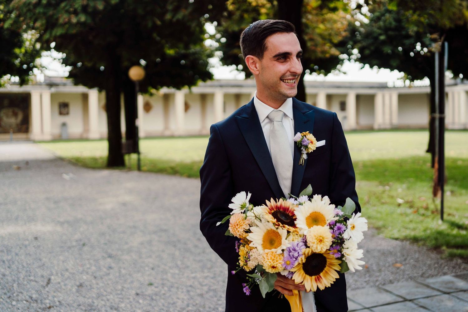 Groom in navy suit holding colorful sunflower wedding bouquet outdoors at elegant venue.
