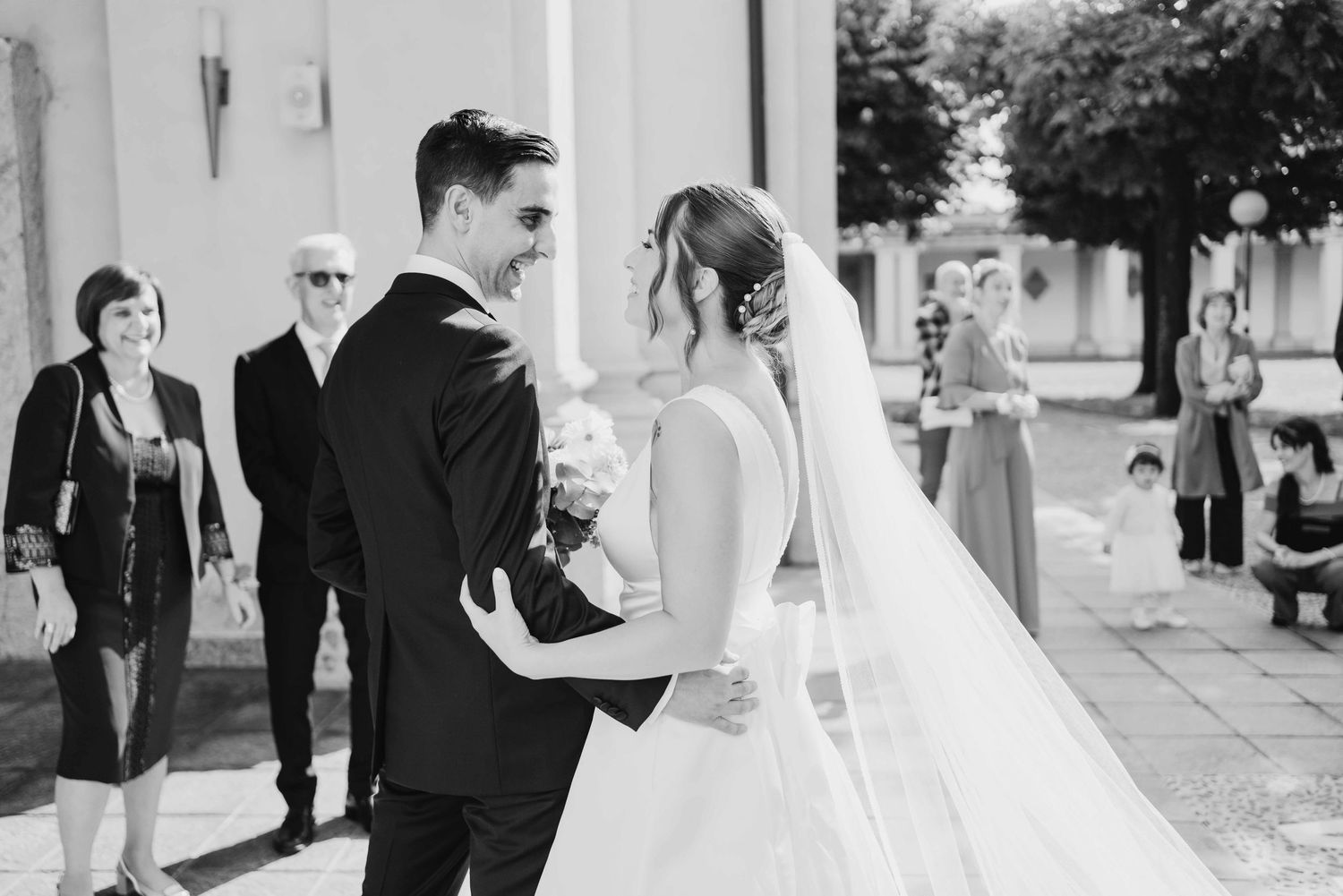 Black and white wedding photo of a smiling couple embracing outdoors, with guests watching in the background.
