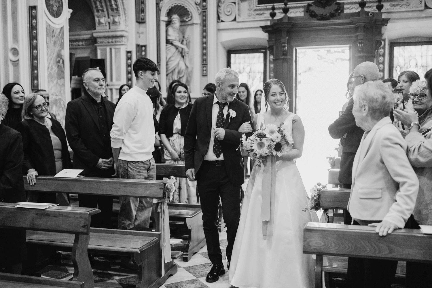 Bride walks down church aisle with escort in black and white wedding ceremony photo.