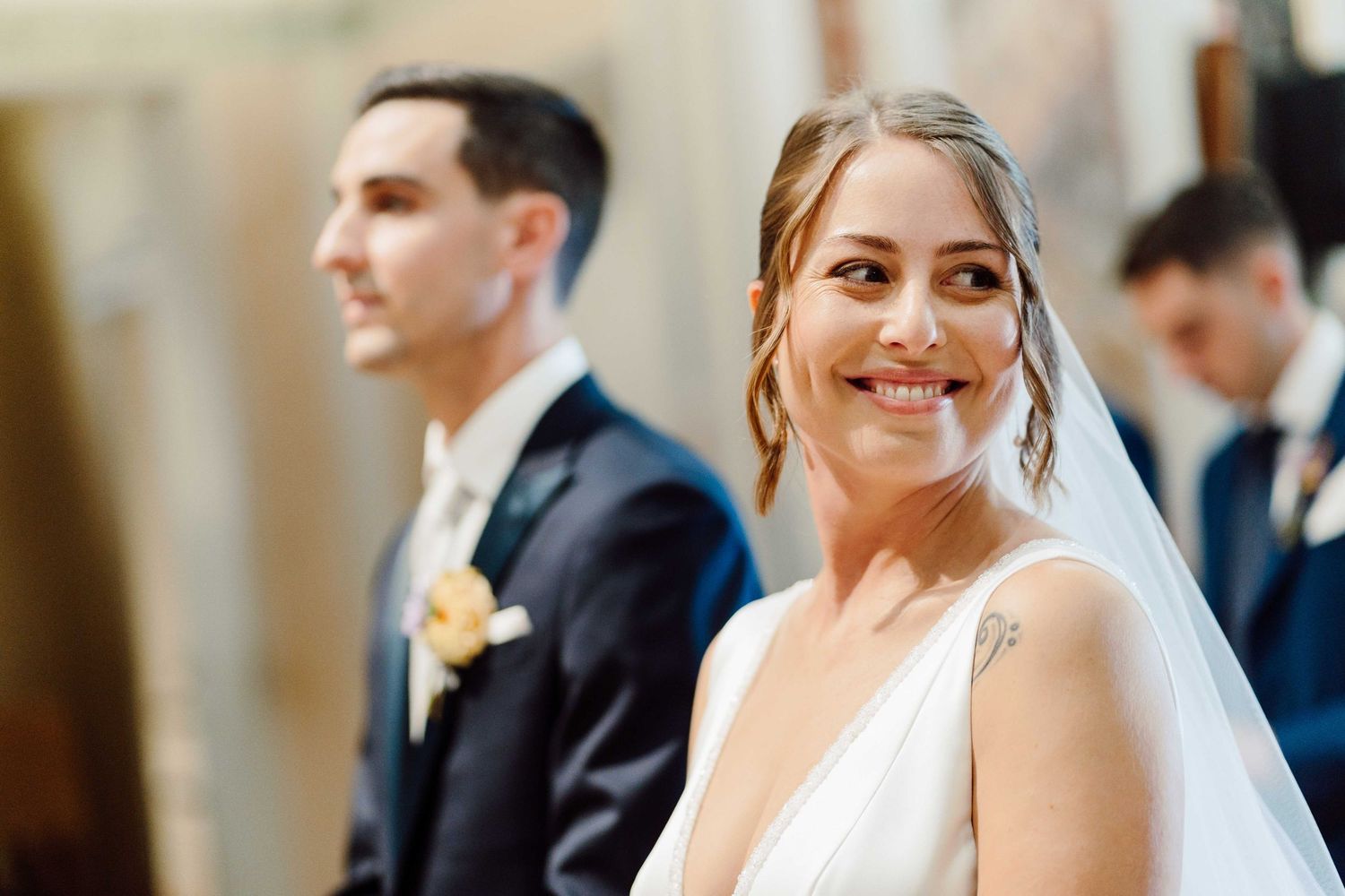 Smiling bride in white gown and veil stands before groom in dark suit at elegant wedding ceremony.