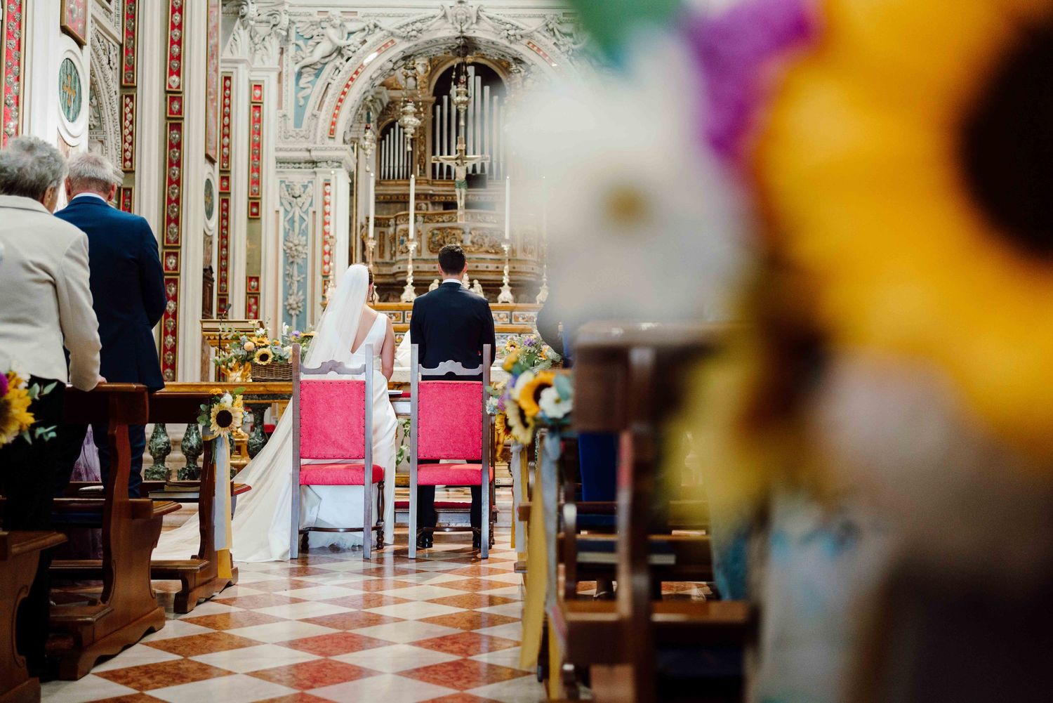A bride and groom kneel at the altar of an ornate church decorated with sunflowers during their wedding ceremony.