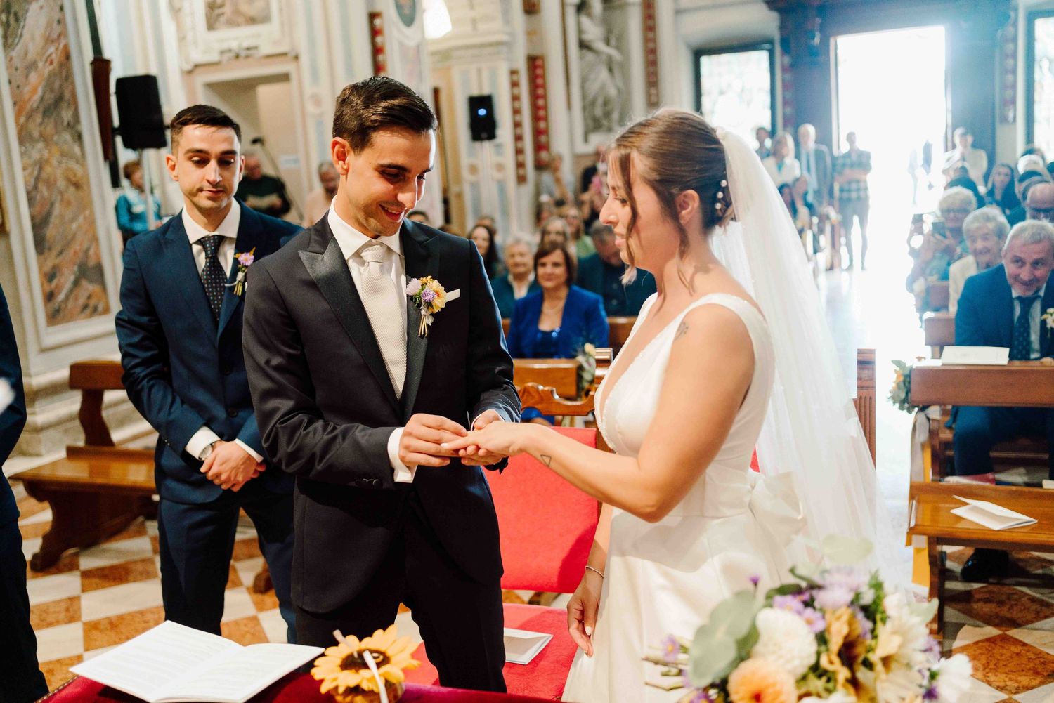 Bride and groom exchanging rings during a church wedding ceremony, with guests seated in pews in the background.