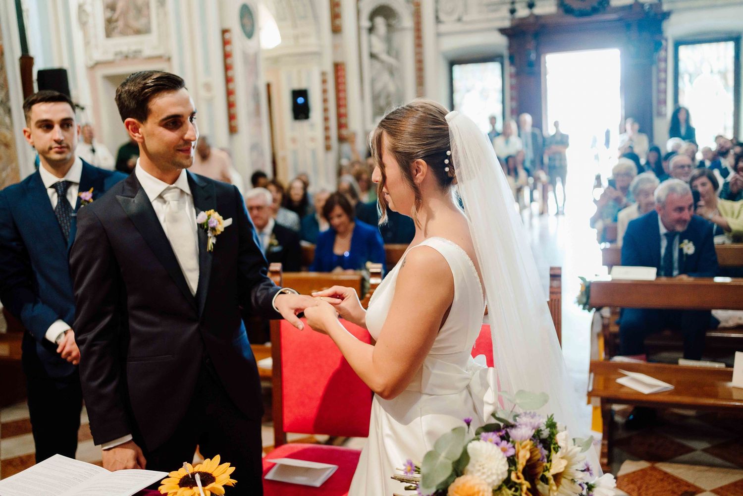 Bride placing wedding ring on groom's finger during church ceremony with guests seated in pews.