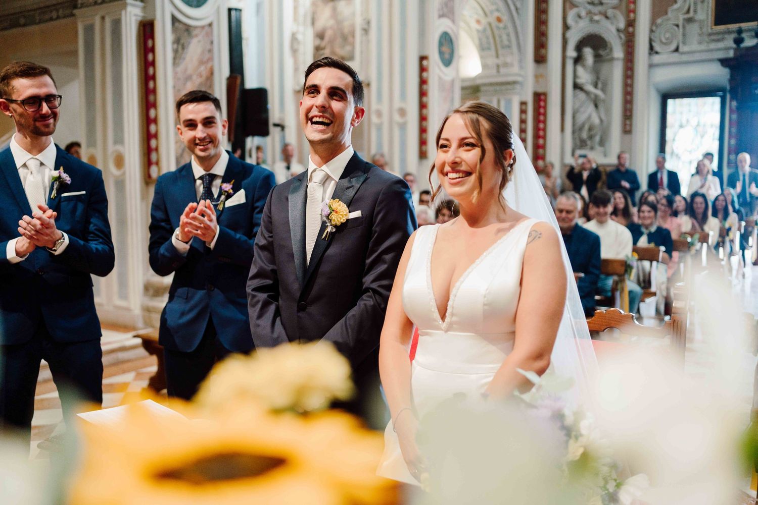 Joyful bride and groom laughing at wedding ceremony altar with groomsmen clapping, sunflowers in foreground.