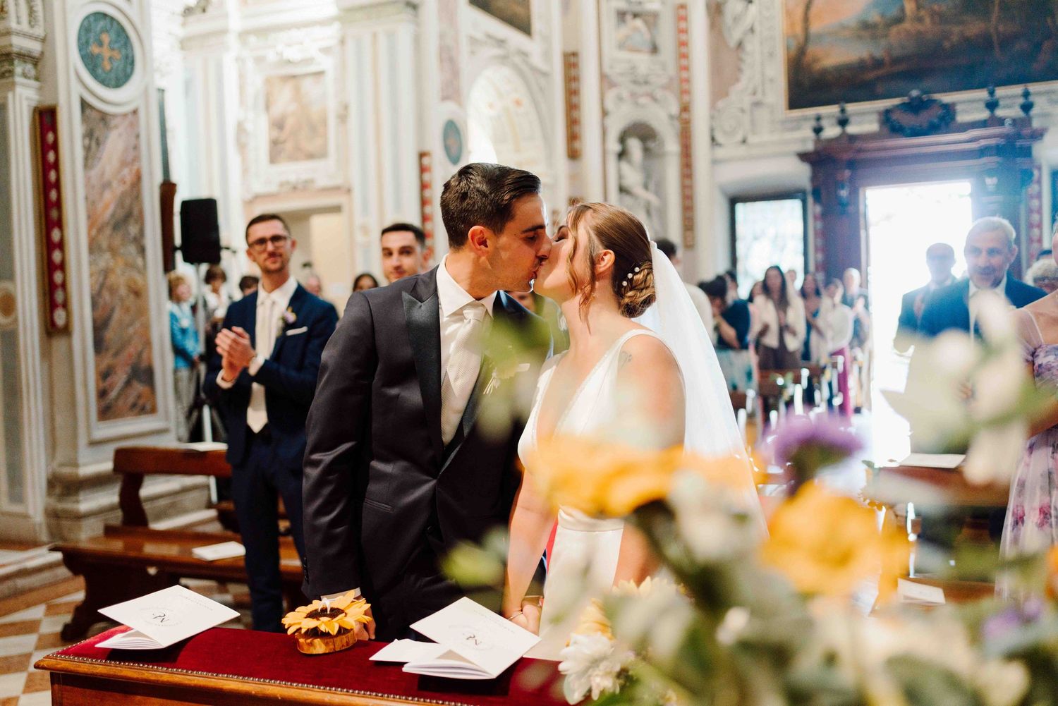 Bride and groom share first kiss at elegant church wedding ceremony with guests applauding in ornate Italian chapel.