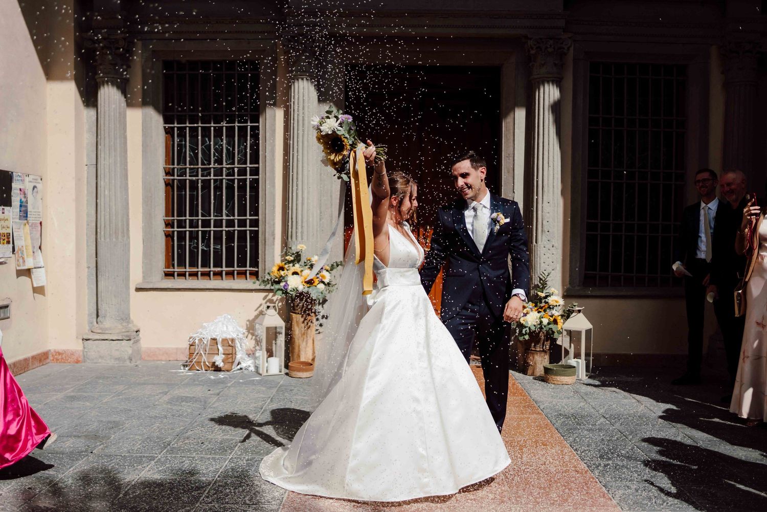 Newlywed couple celebrating outside church, bride in white gown holding bouquet as confetti falls.