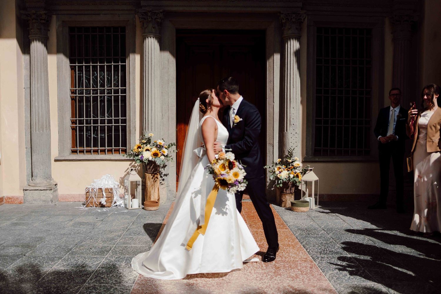 Bride and groom share a kiss outside a sunlit church, bride holding a sunflower bouquet with yellow ribbon.