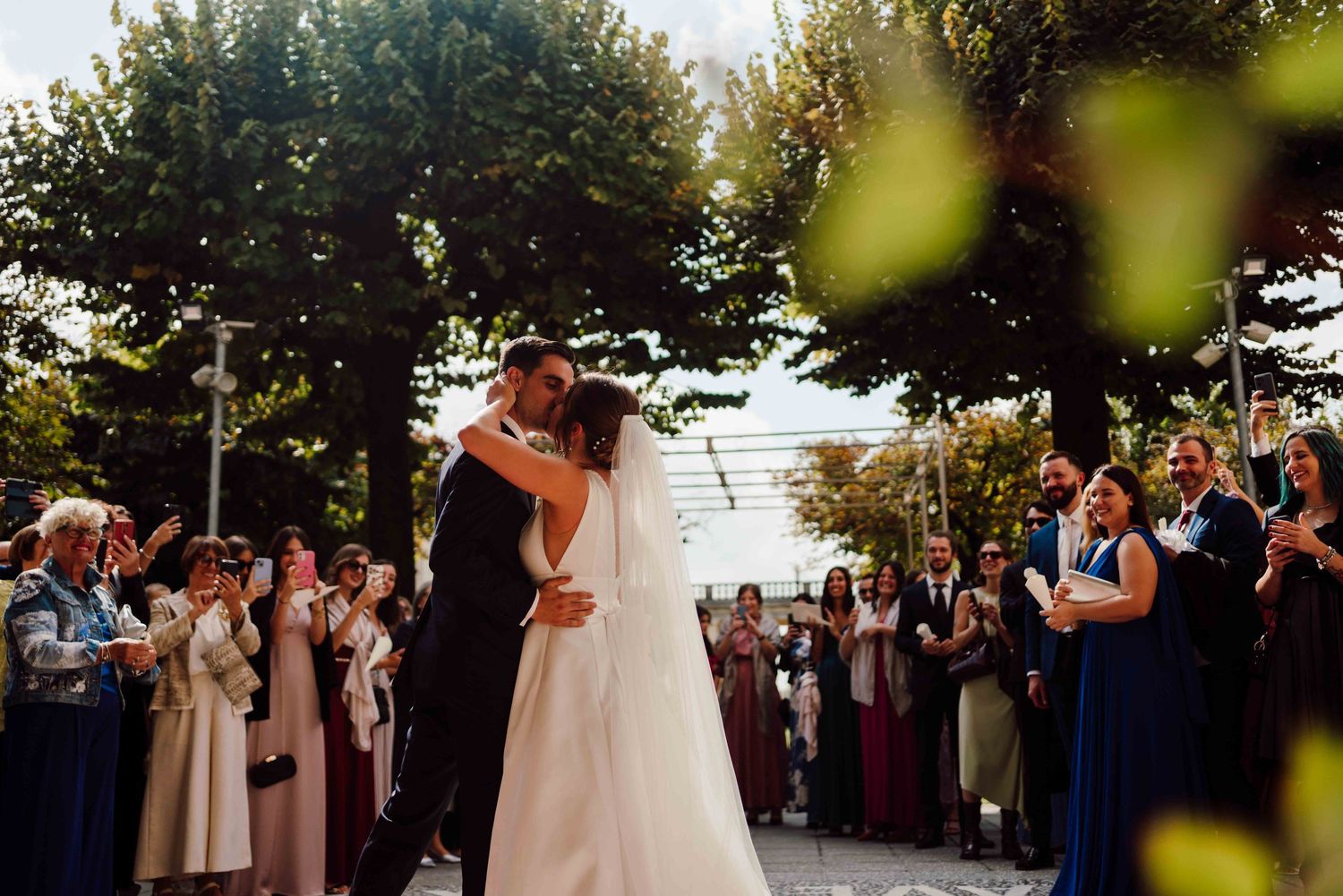 Bride and groom share first dance kiss surrounded by cheering guests outdoors at elegant wedding ceremony.