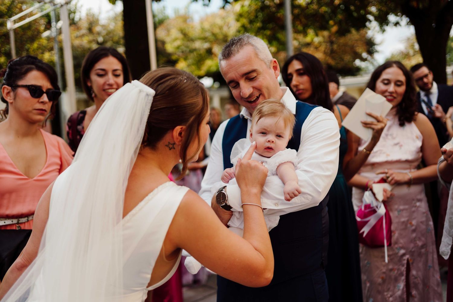 Bride and groom holding a baby at outdoor wedding reception with guests in background.
